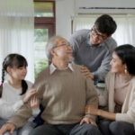 Asian elderly senior male grandpa sitting at sofa couch at home living room on quarantine in concept healthcare, chronic health issue or low back pain in retired older people with family.